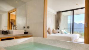 A modern hotel room with a large bed, a bathtub, and a view of mountains through a large window. - Botanica Khao Yai (Pak Chong)