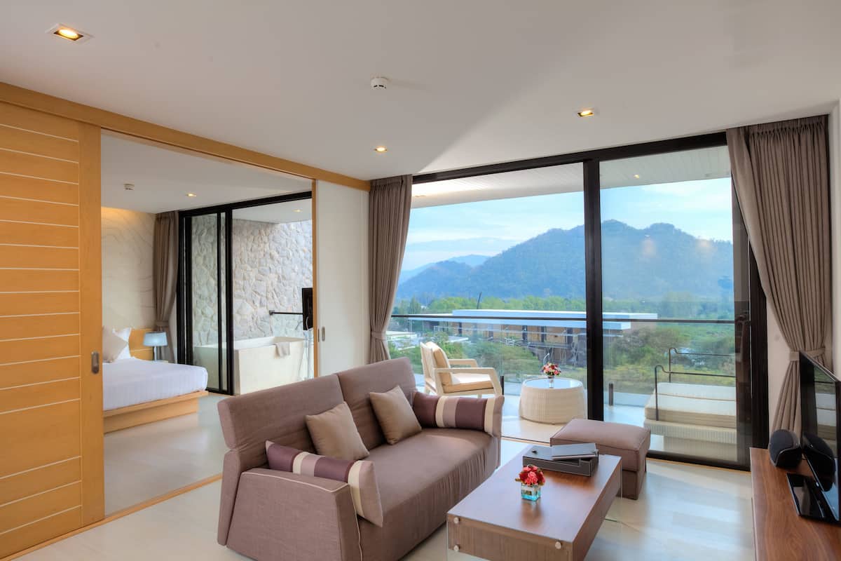 A modern hotel room with a large bed, a sofa, a coffee table, and a balcony with a mountain view.