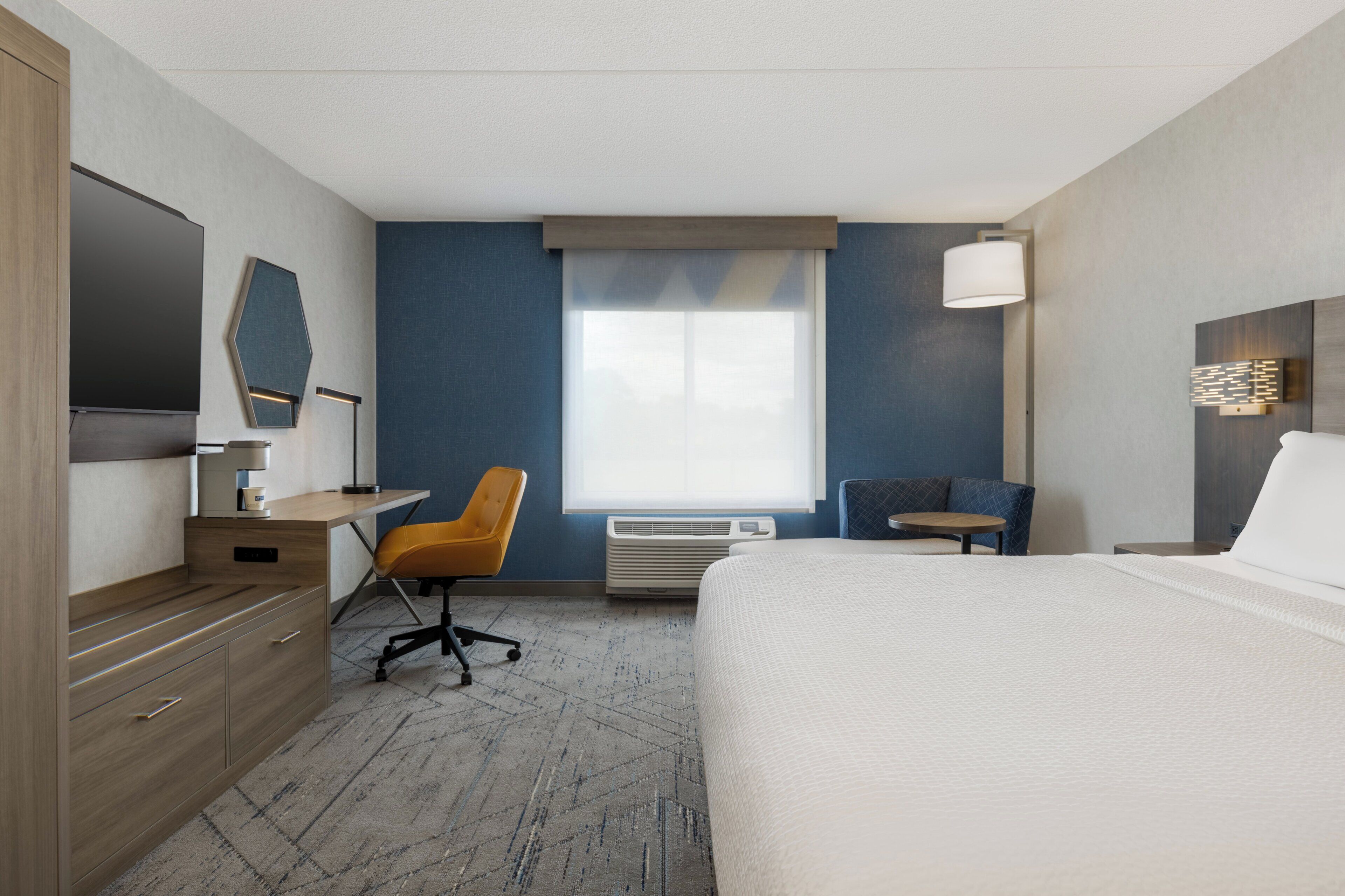 Une chambre d’hôtel avec un grand lit, un bureau avec une chaise, une télévision et une fenêtre avec des stores.