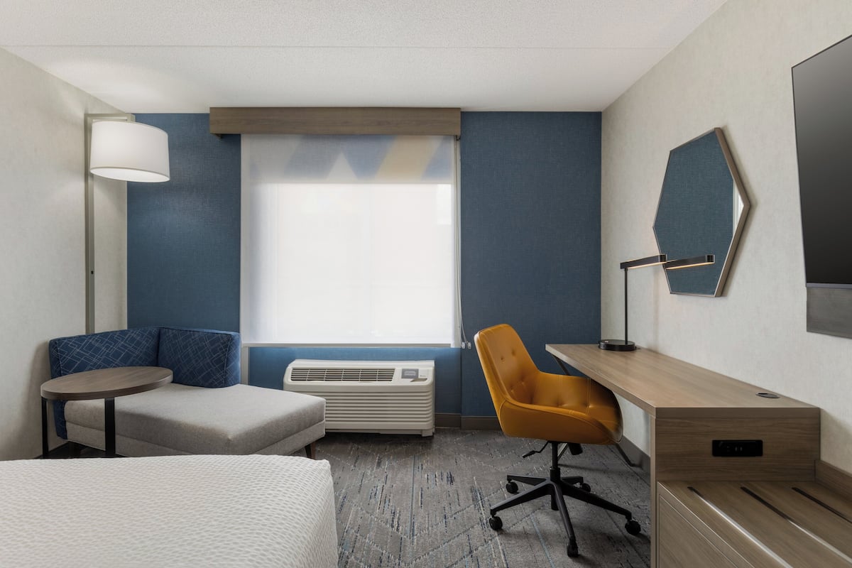 A hotel room with a bed, a desk, a chair, a lamp, a heater, and a large window.