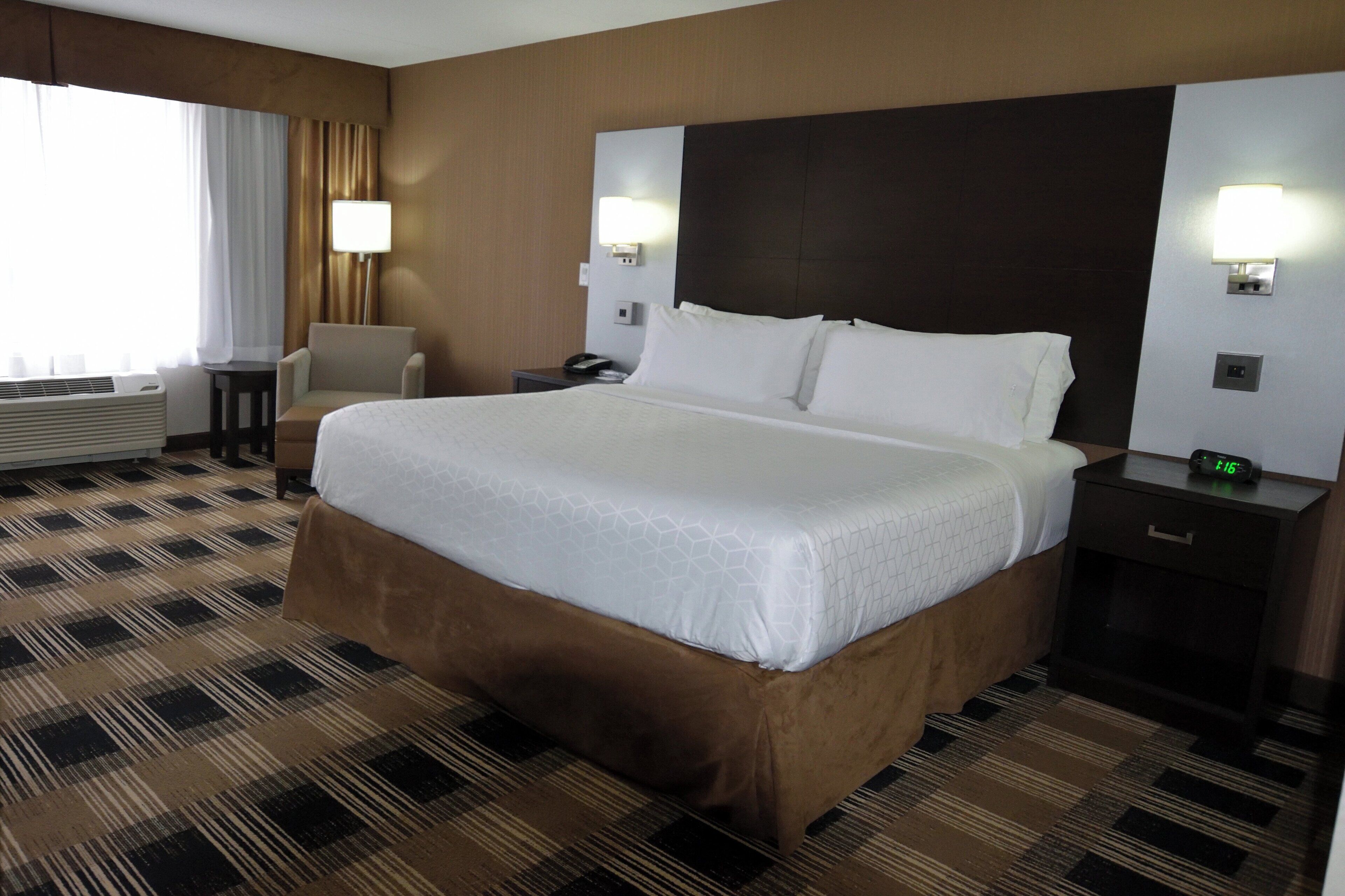 A hotel room with a large bed, a chair, a bedside table, a lamp, and a clock.