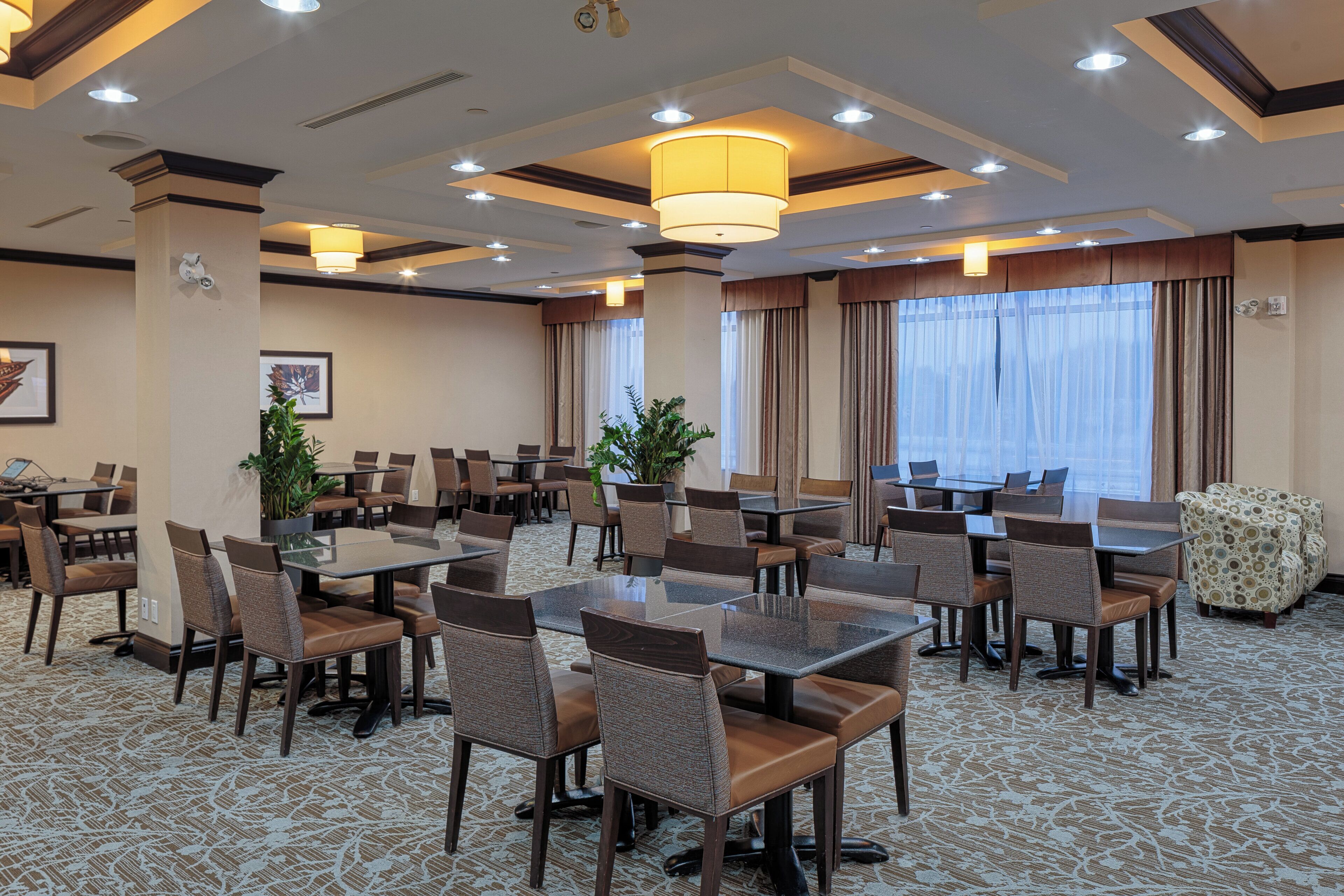 Un salón de conferencias de hotel con varias mesas y sillas, ventanas amplias con cortinas e iluminación empotrada en el techo.
