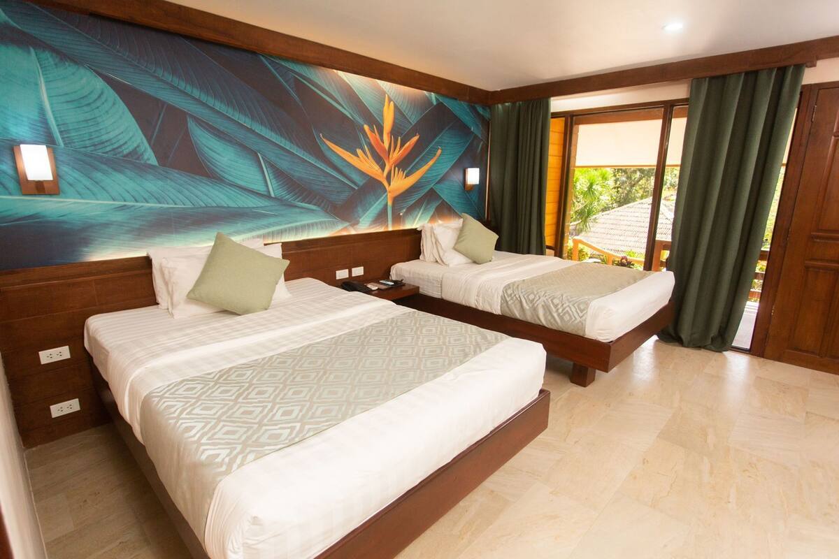 A hotel room with two beds, a large tropical mural, and a view of greenery outside.
