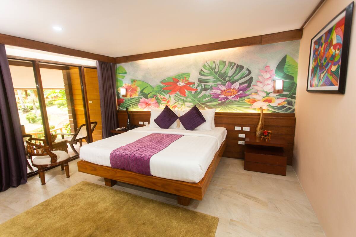 A hotel room with a bed, wooden headboard, a mural of tropical flowers, a nightstand, a chair, and a view of the outdoors.