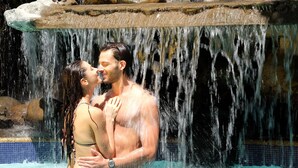 Um casal se abraçando em uma piscina com uma cachoeira ao fundo.