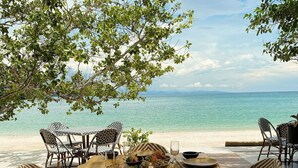 Mesa de jantar posta com diversos pratos e bebidas, com vista para uma praia e um mar tranquilo, além de montanhas ao longe.