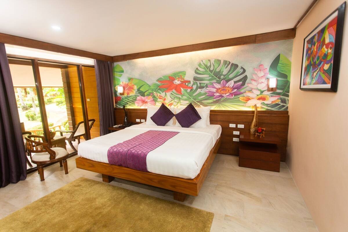 A bedroom with a bed, wooden headboard, a floral mural, a nightstand, a chair, and a view of the outdoors.