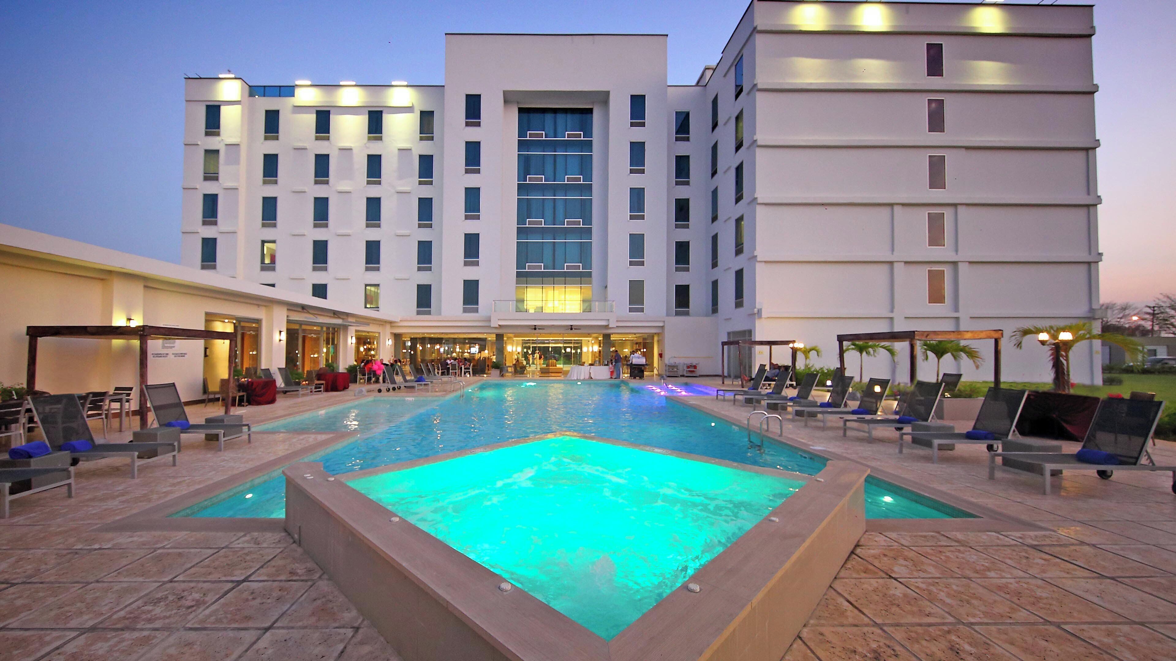 A hotel with a swimming pool, lounge chairs, and a building with multiple floors.