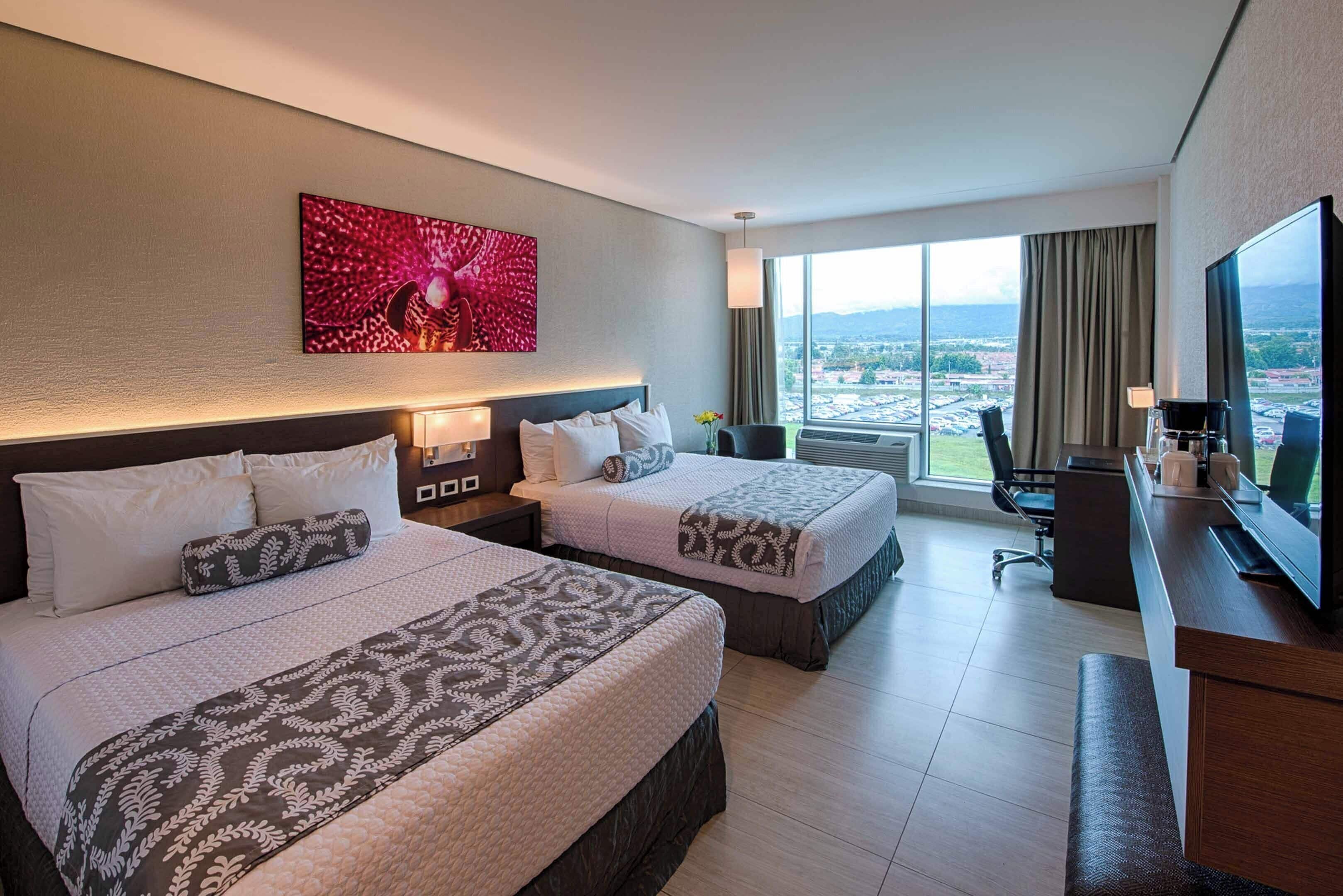 A hotel room with two beds, a desk, a TV, and a large window with a city view.