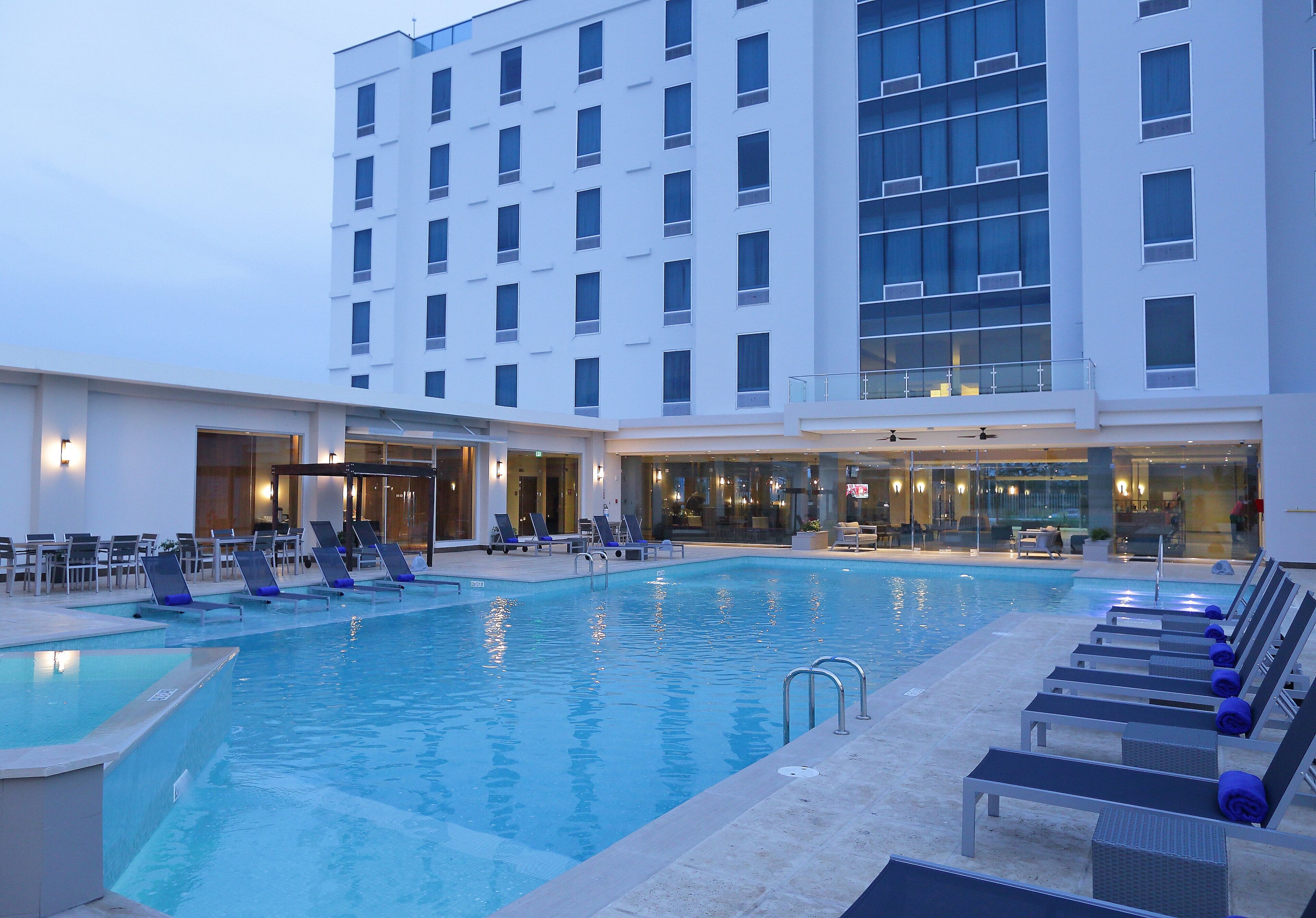 A hotel pool area with lounge chairs and a building with multiple floors and large windows.