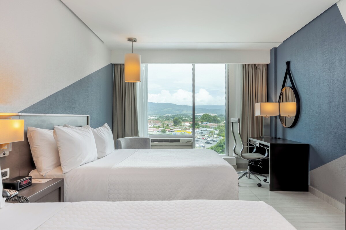 Habitación de hotel con cama, mesita de noche, lámpara, escritorio, silla y un gran ventanal con vistas a la ciudad.