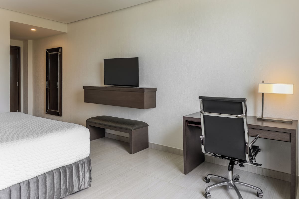 Habitación de hotel con cama, escritorio con silla, televisor en estante y lámpara de noche.