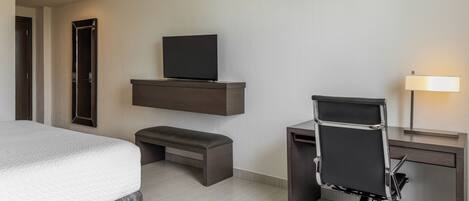 A hotel room with a bed, a desk with a chair, a TV on a shelf, and a bedside lamp.