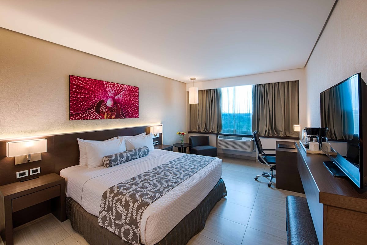 Habitación de hotel con una cama grande, un escritorio con computadora, un televisor y una ventana con cortinas.
