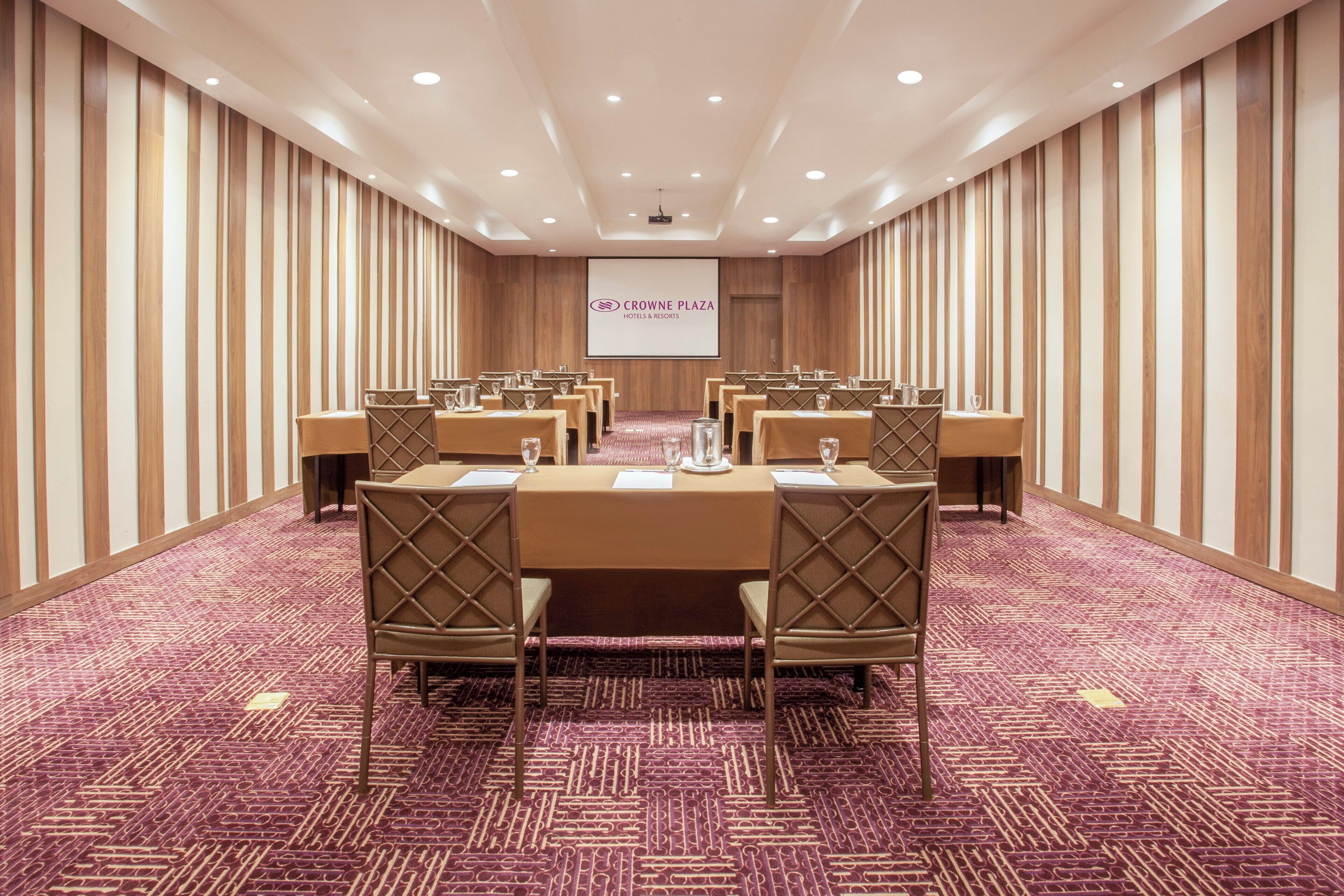 Un salón de conferencias con una larga mesa, sillas y una pantalla de proyección que muestra 'Crown Plaza Hotels & Resorts'.