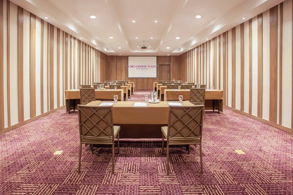 Un salón de conferencias con una larga mesa, sillas y una pantalla de proyección que muestra 'Crown Plaza Hotels & Resorts'.