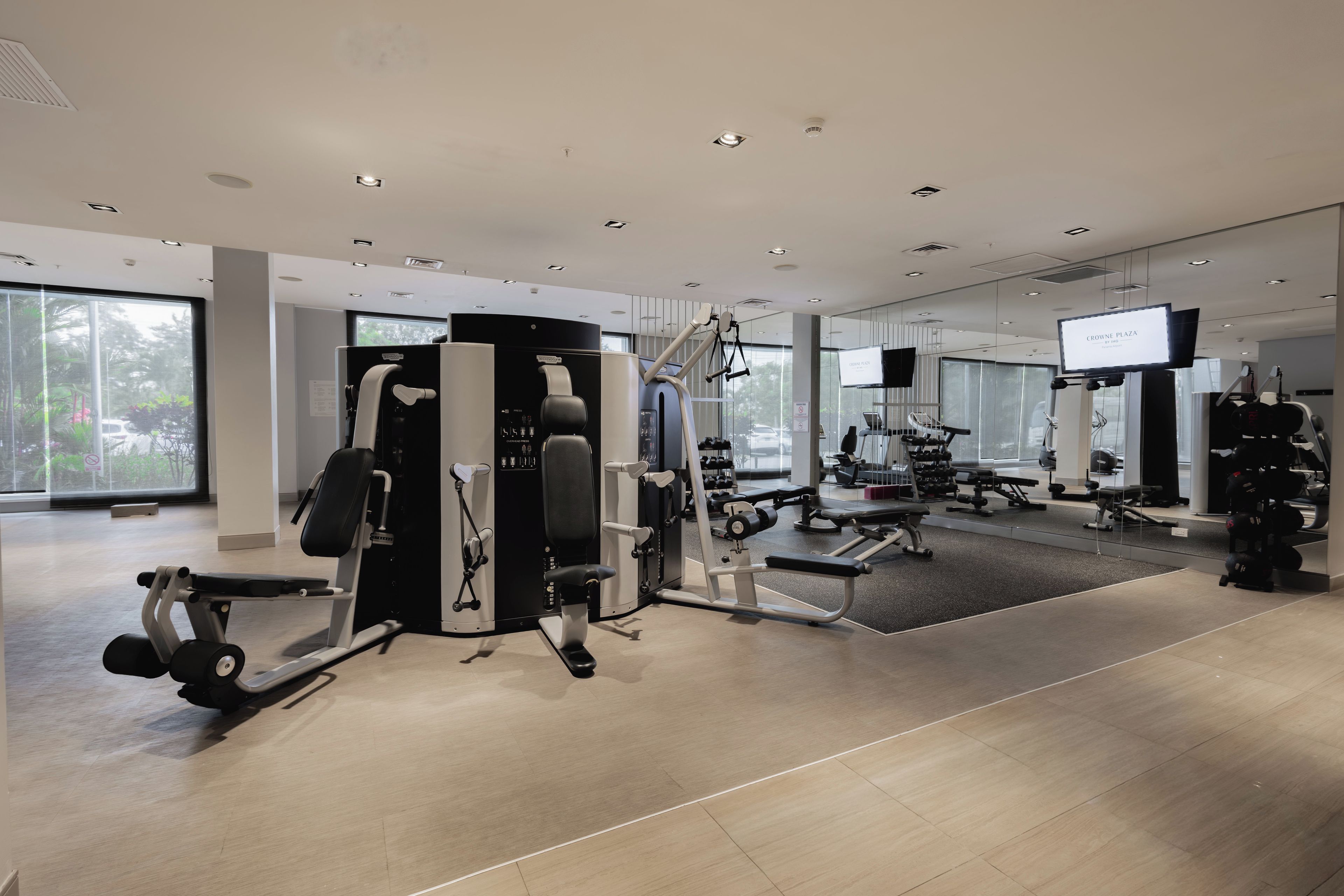 A modern gym with various exercise equipment, large windows, and a television screen.