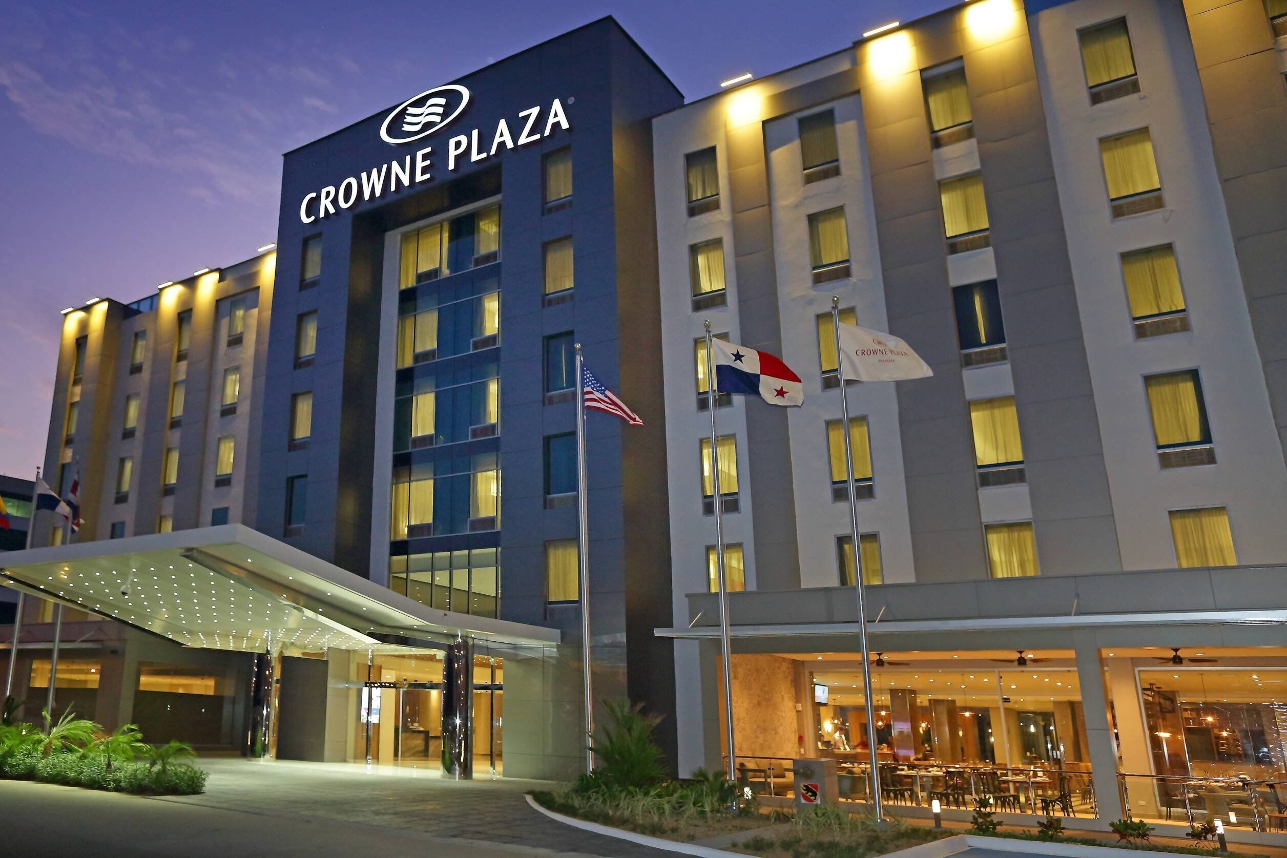 Un hotel con un llamativo letrero 'CROWNE PLAZA', varias banderas y una entrada bien iluminada.
