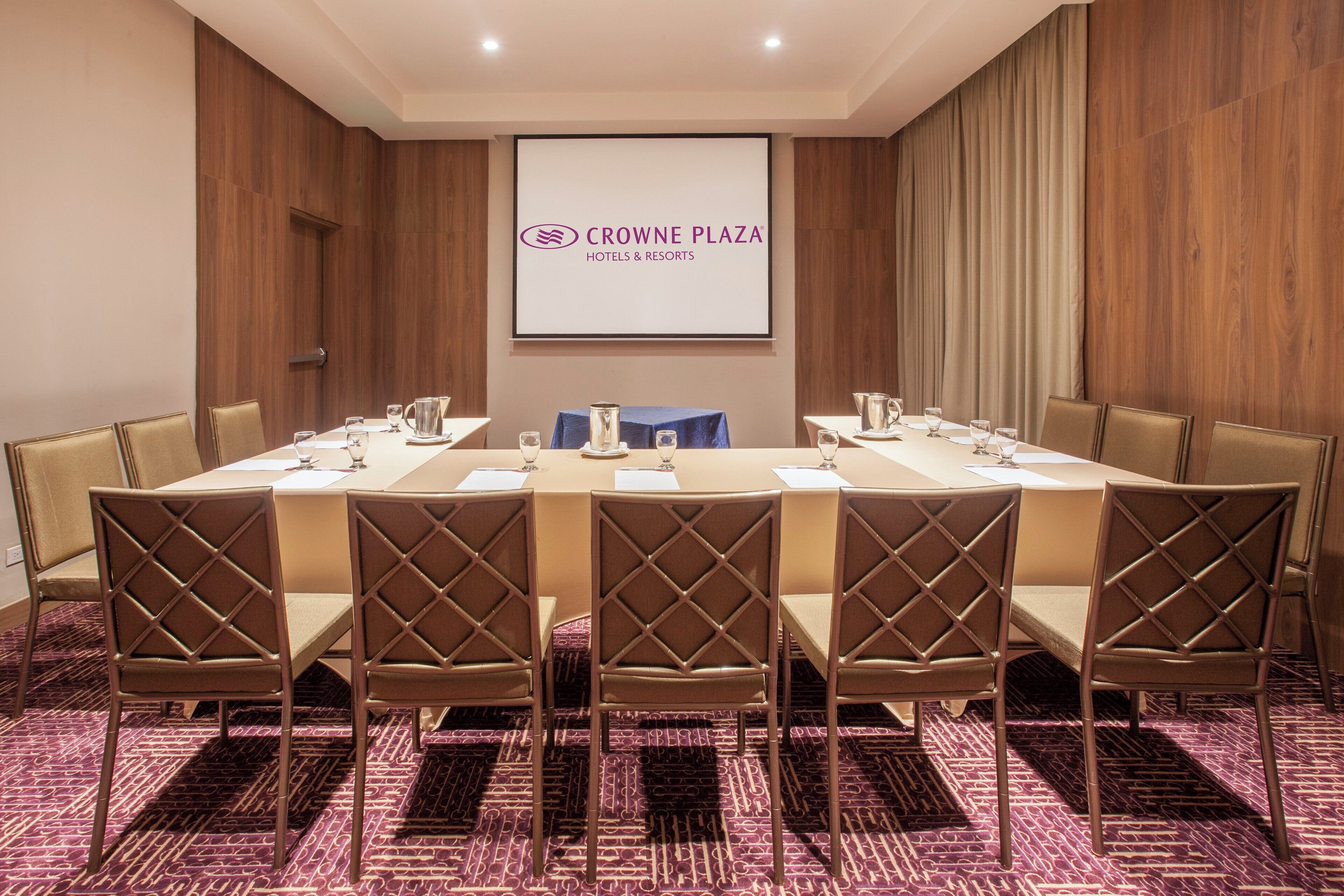 Une salle de conférence avec une longue table, six chaises et un écran affichant le logo de « CROWN PLAZA HOTELS & RESORTS ».