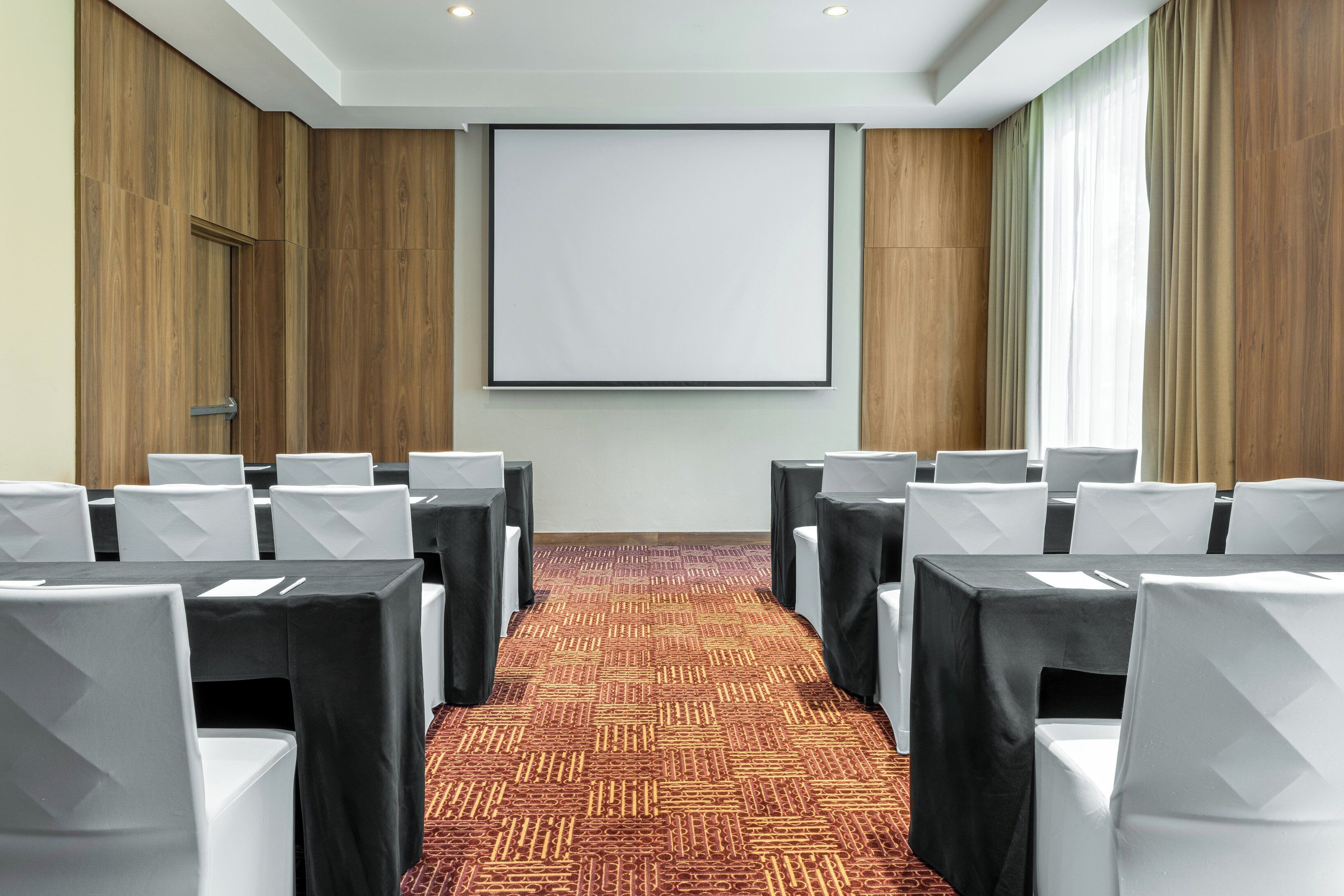 Un salón de conferencias con filas de sillas blancas, manteles negros y una pantalla grande en blanco en la pared.