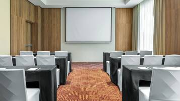 Un salón de conferencias con filas de sillas blancas, manteles negros y una pantalla grande en blanco en la pared.