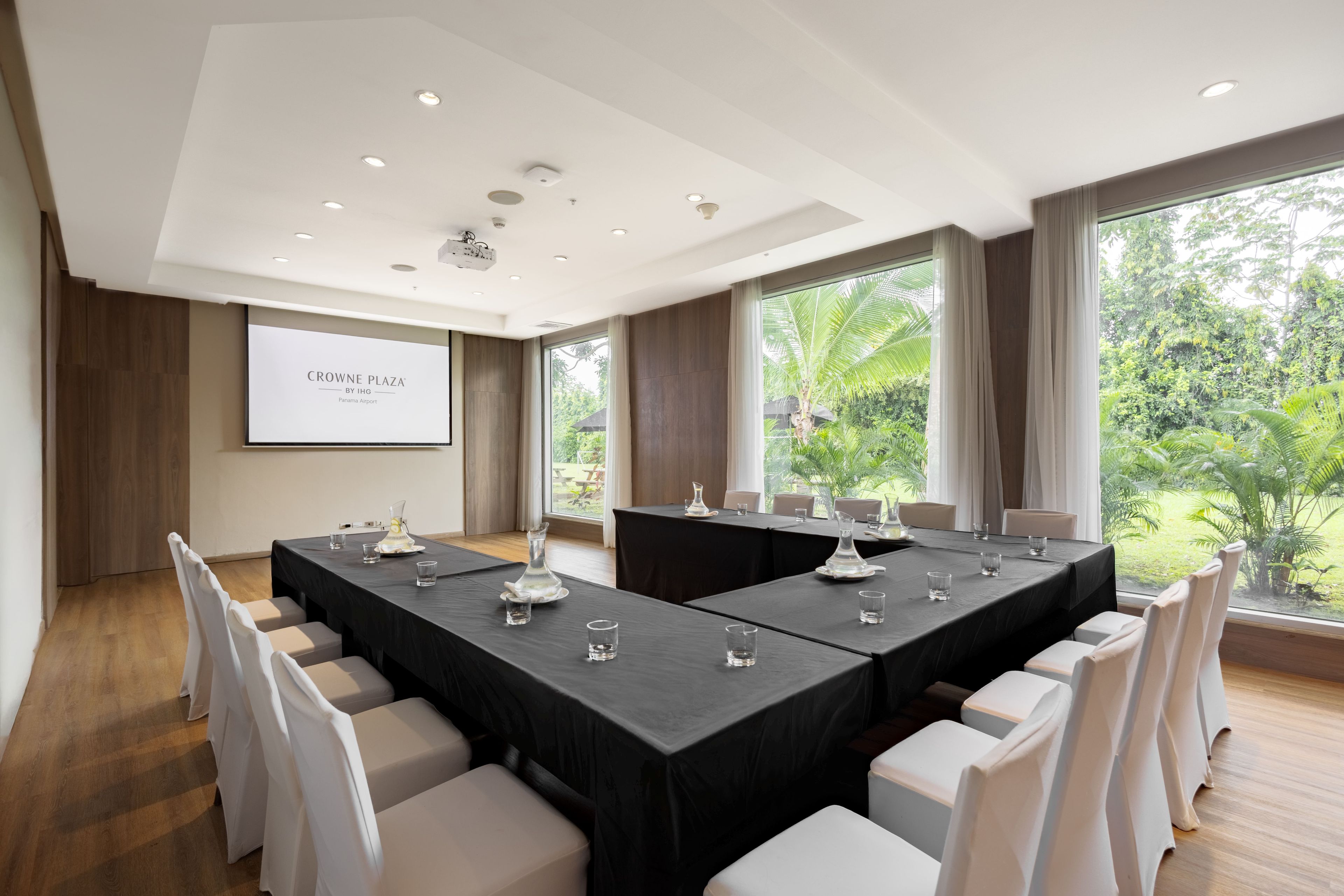 Un salón de conferencias con pantalla de proyector, ventanas amplias y vistas a zonas verdes al exterior.