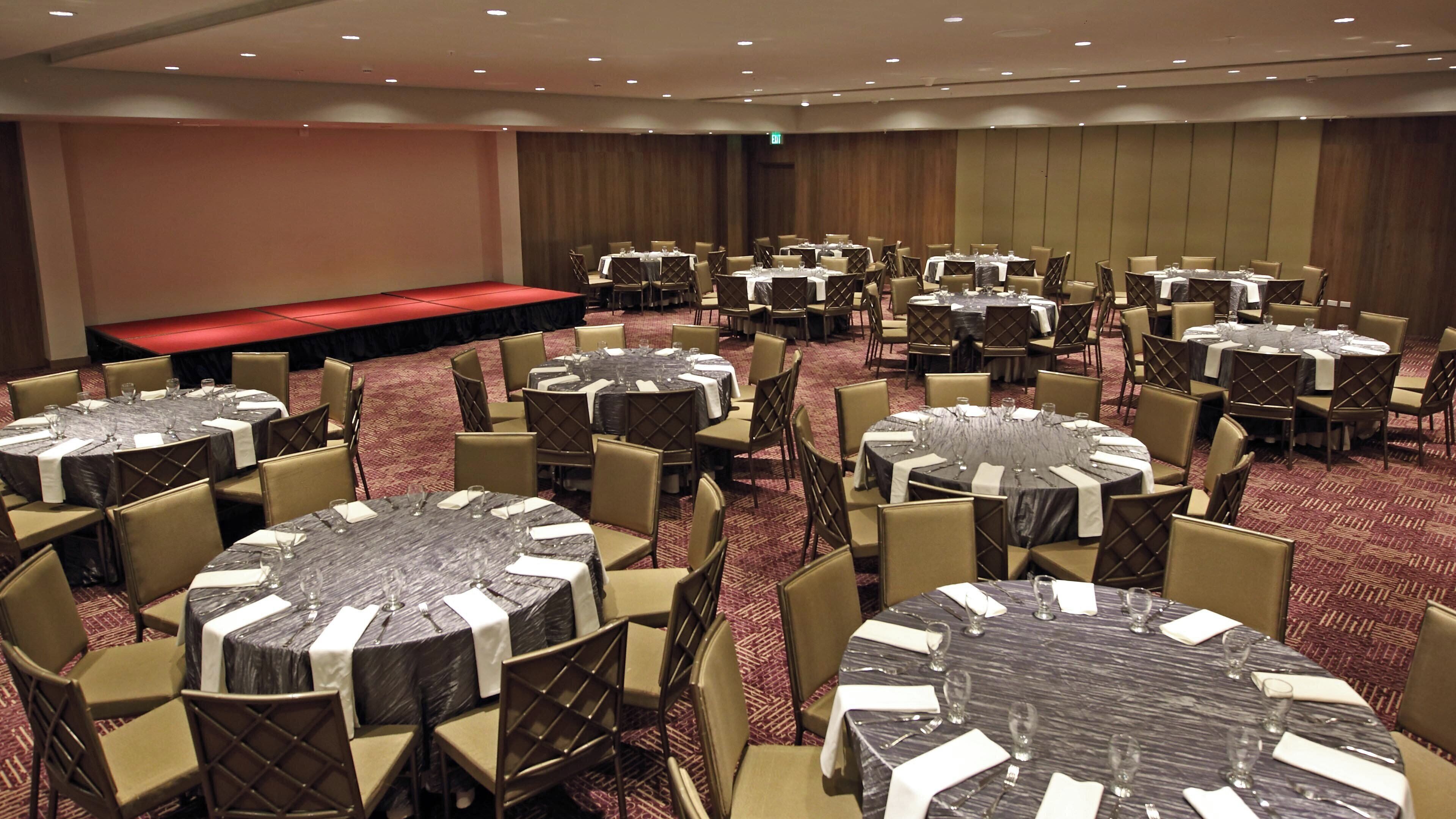A conference room with multiple round tables, each set with chairs and tablecloths, and a stage area with a red carpet.