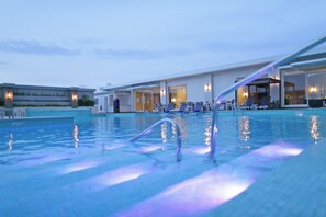 Una piscina con superficie de agua cristalina, una zona junto a la piscina con sillones de descanso y un edificio con grandes ventanales y puertas.