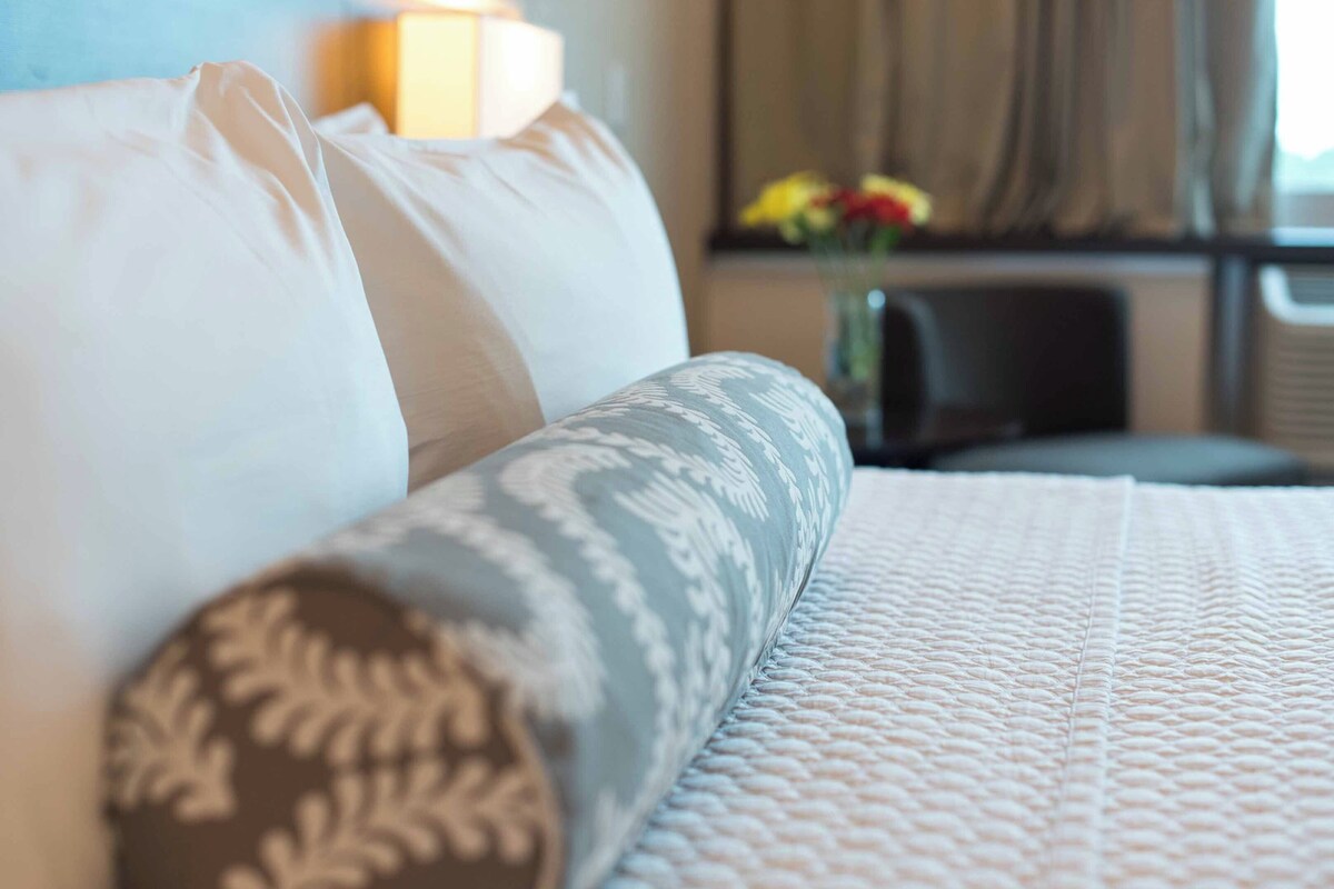 Una habitación de hotel con una cama, almohadas, una mesita de noche y un jarrón con flores.