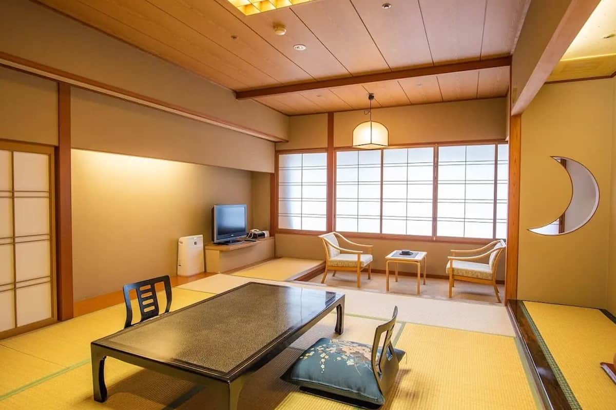 A traditional Japanese room with tatami flooring, a low table, chairs, a television, and a large window with a view.