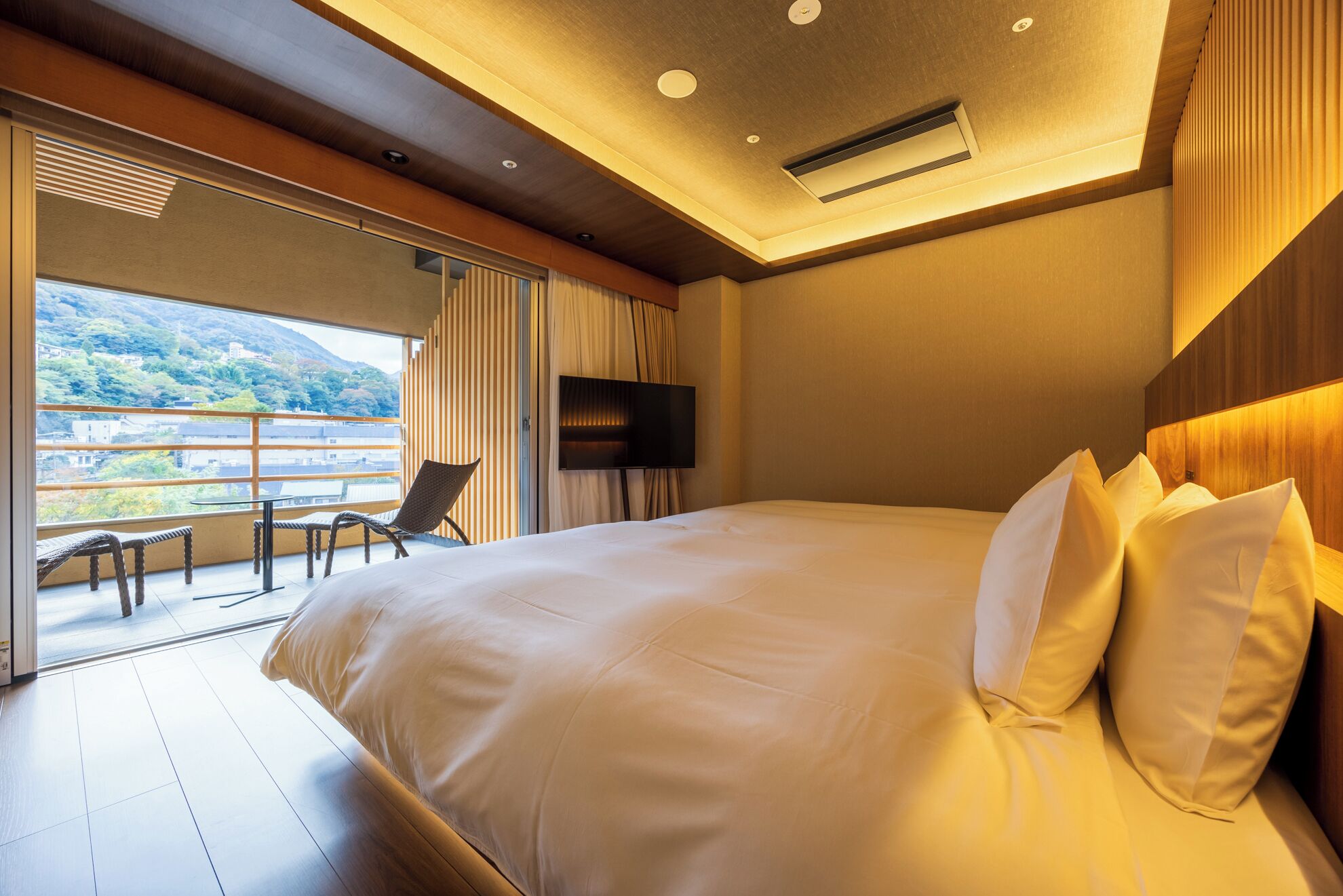 Ein modernes Hotelzimmer mit einem großen Bett, einem flachbildfernseher an der Wand und einem Balkon mit Blick auf die Stadt und die Hügel.