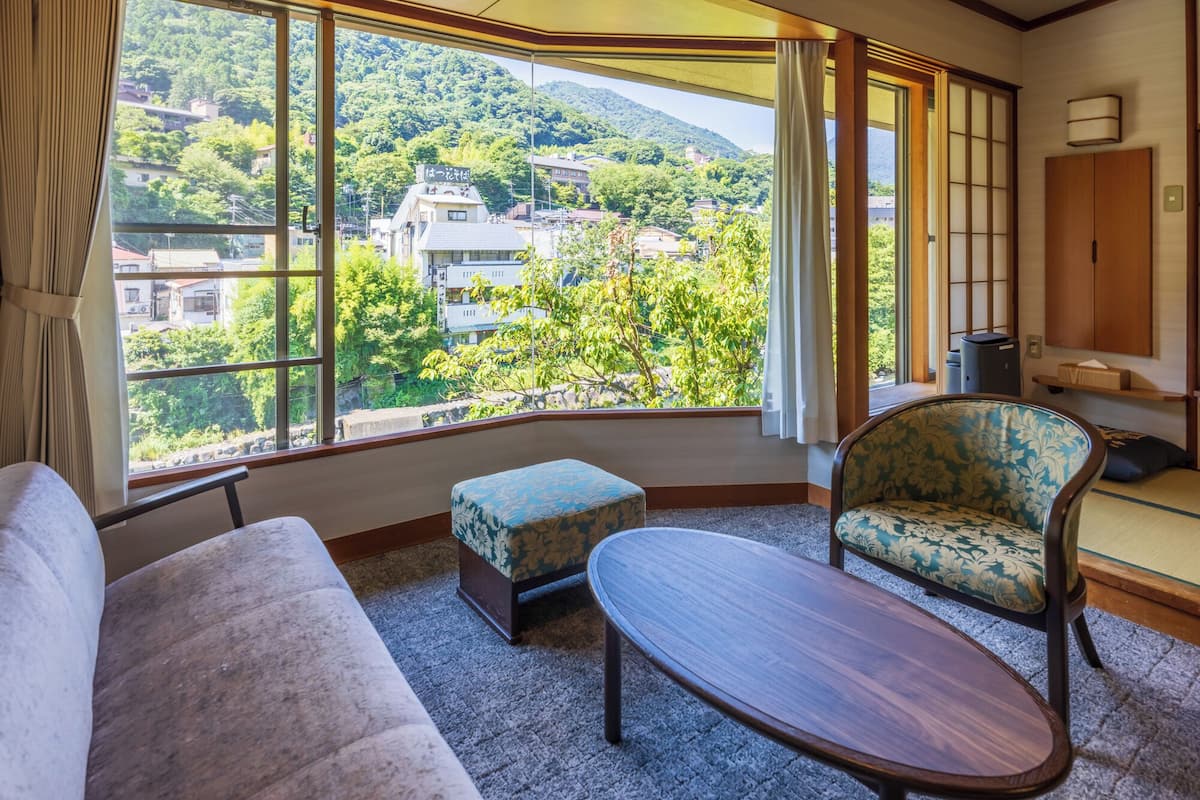 A room with a large window offering a view of a hillside with buildings. There is a sofa, a chair, and a small table.