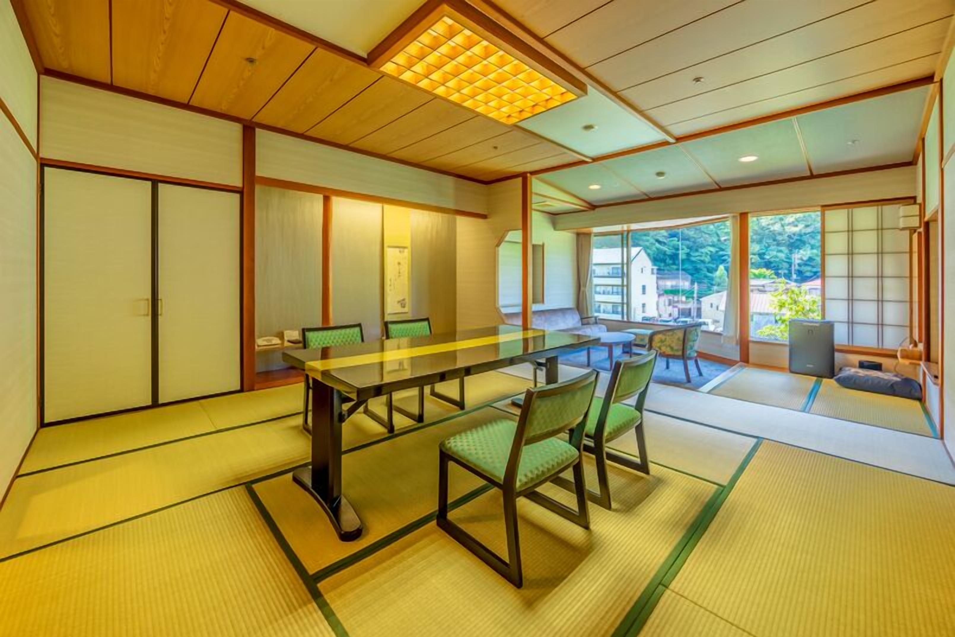 Um quarto japonês tradicional com tatames, uma mesa baixa com cadeiras, portas deslizantes e vista para um edifício lá fora.