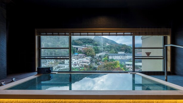 A modern bathroom with a glass-enclosed hot tub, a view of a town and mountains, and a wooden bench. - Hotel Kajikaso (Hakone)