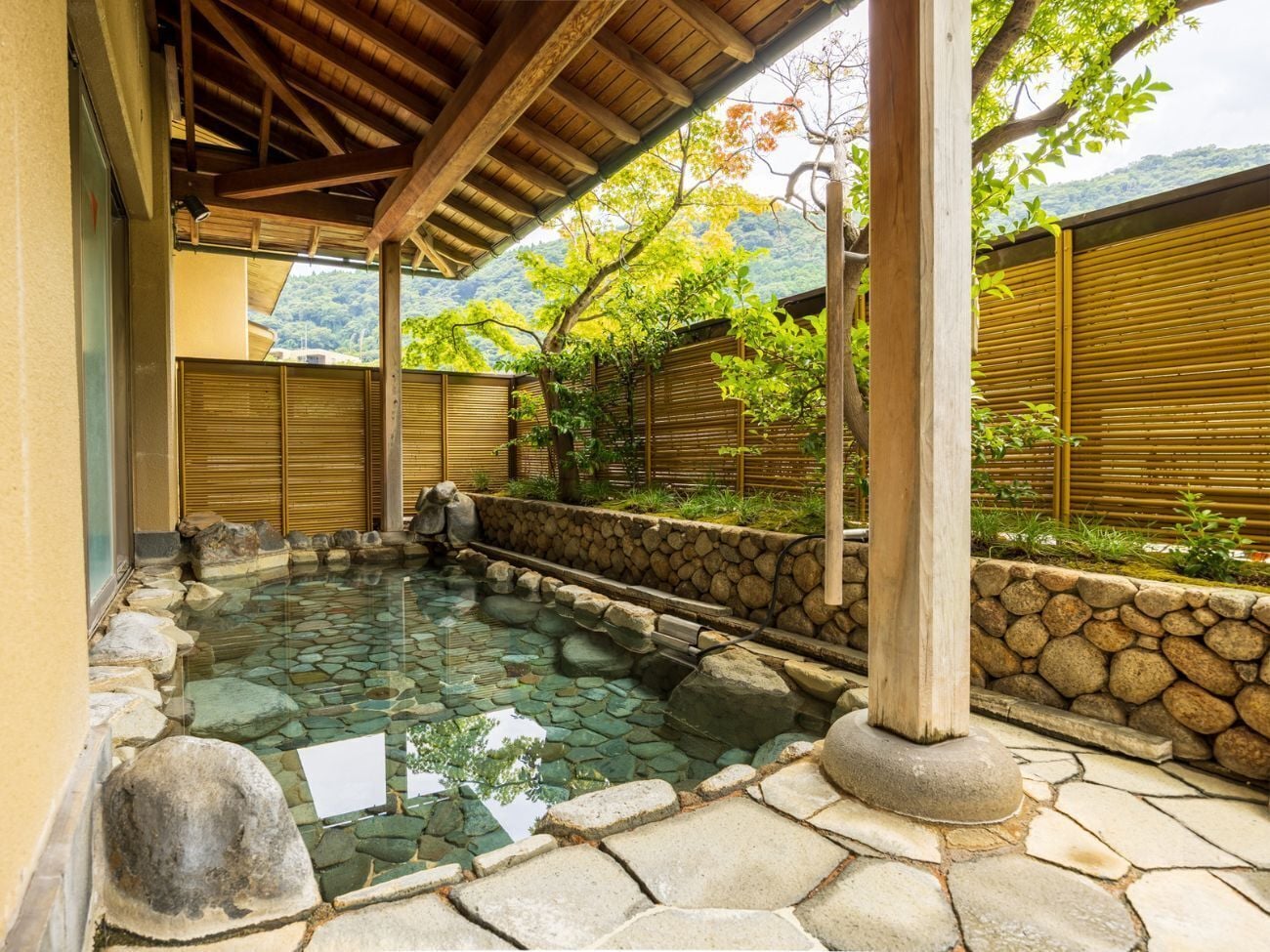 Un onsen japonés tradicional con piscina revestida de piedra, pilares de madera y una cerca de bambú.