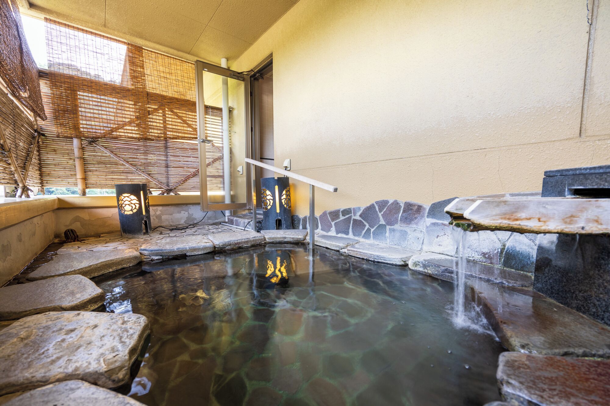 A hot spring area with stone steps leading into the water, a wooden bench, and a bamboo screen.