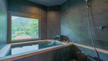 A modern bathroom with a large bathtub, a shower, and a window offering a view of a forested area.
