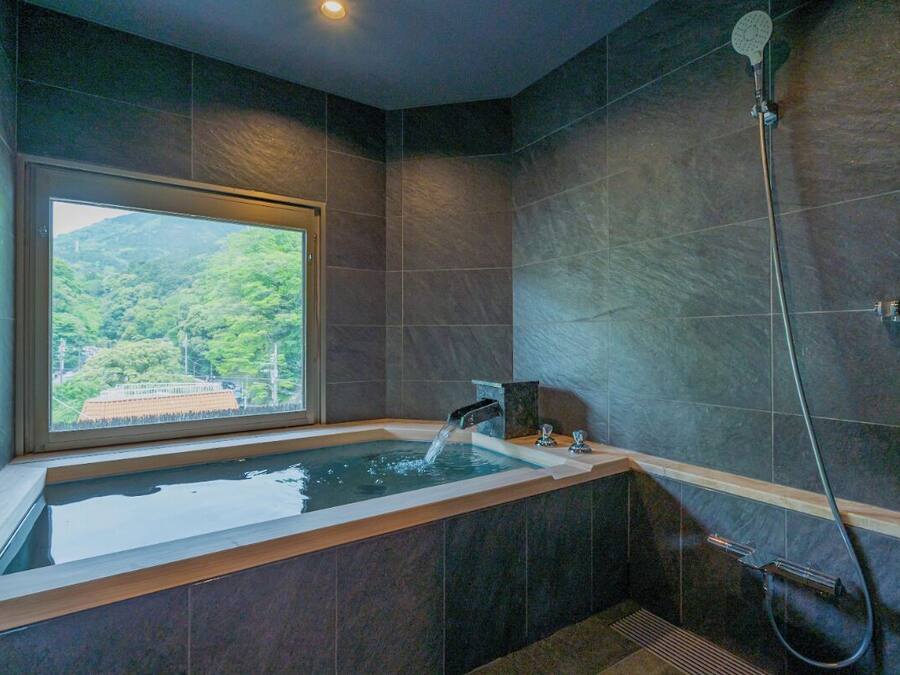 A modern bathroom with a large bathtub, a shower, and a window offering a view of a forested area.