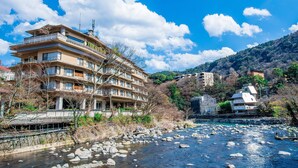 Un hotel de varios pisos con balcones, un río en primer plano y montañas al fondo.