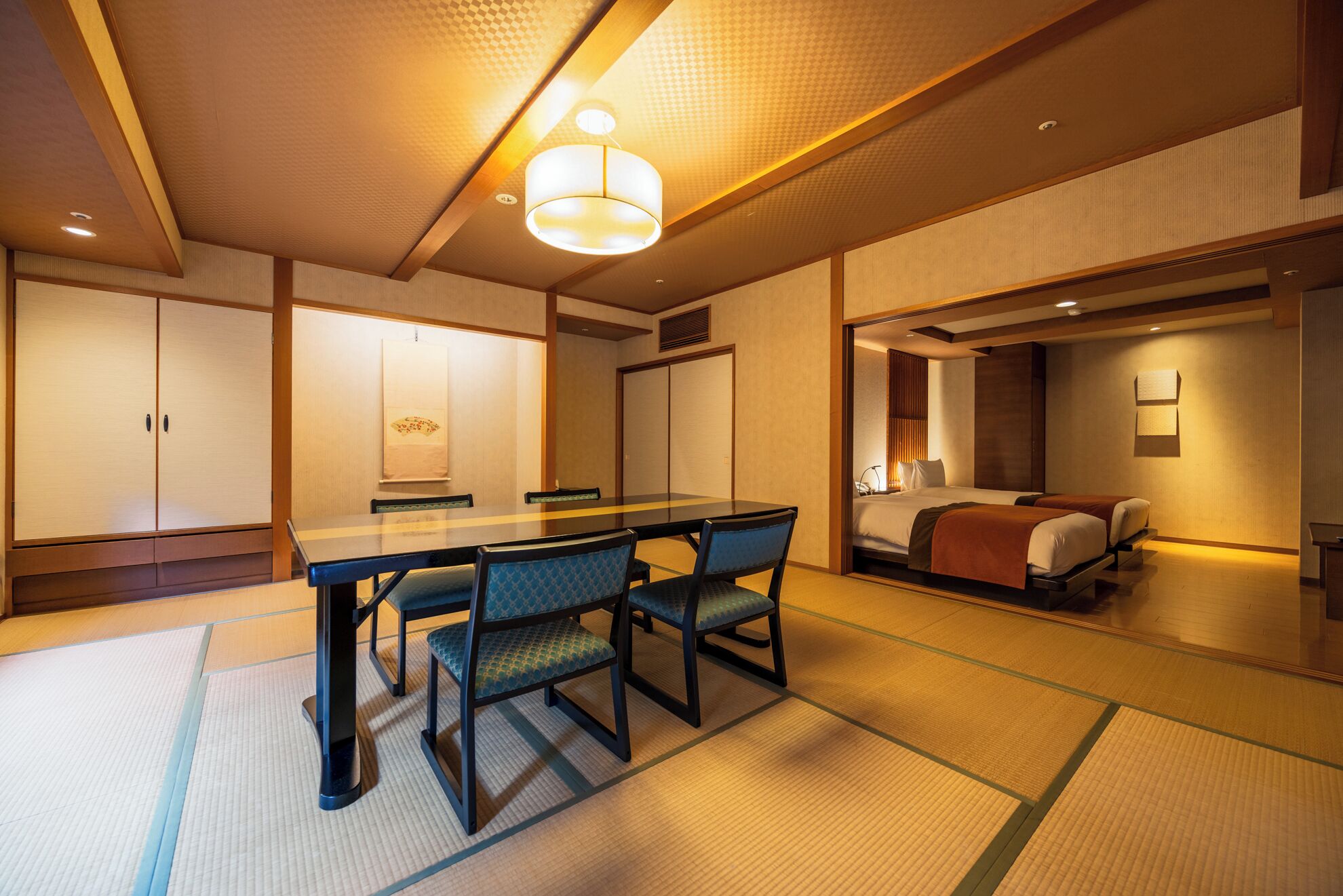 A traditional Japanese-style room with tatami flooring, a low table with chairs, and a bed with a wooden headboard.