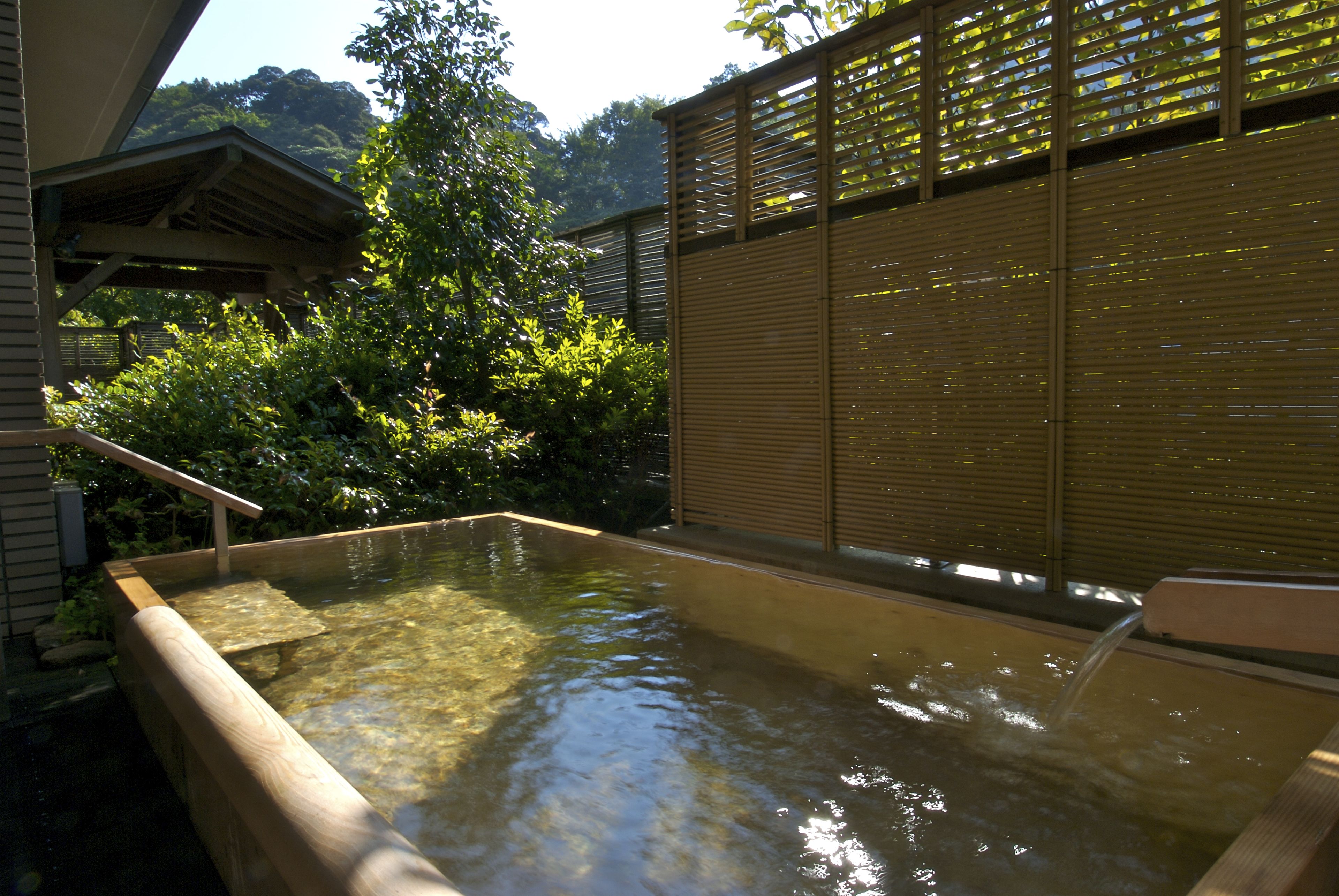 A traditional Japanese onsen with a wooden bath, surrounded by lush greenery and a wooden fence.