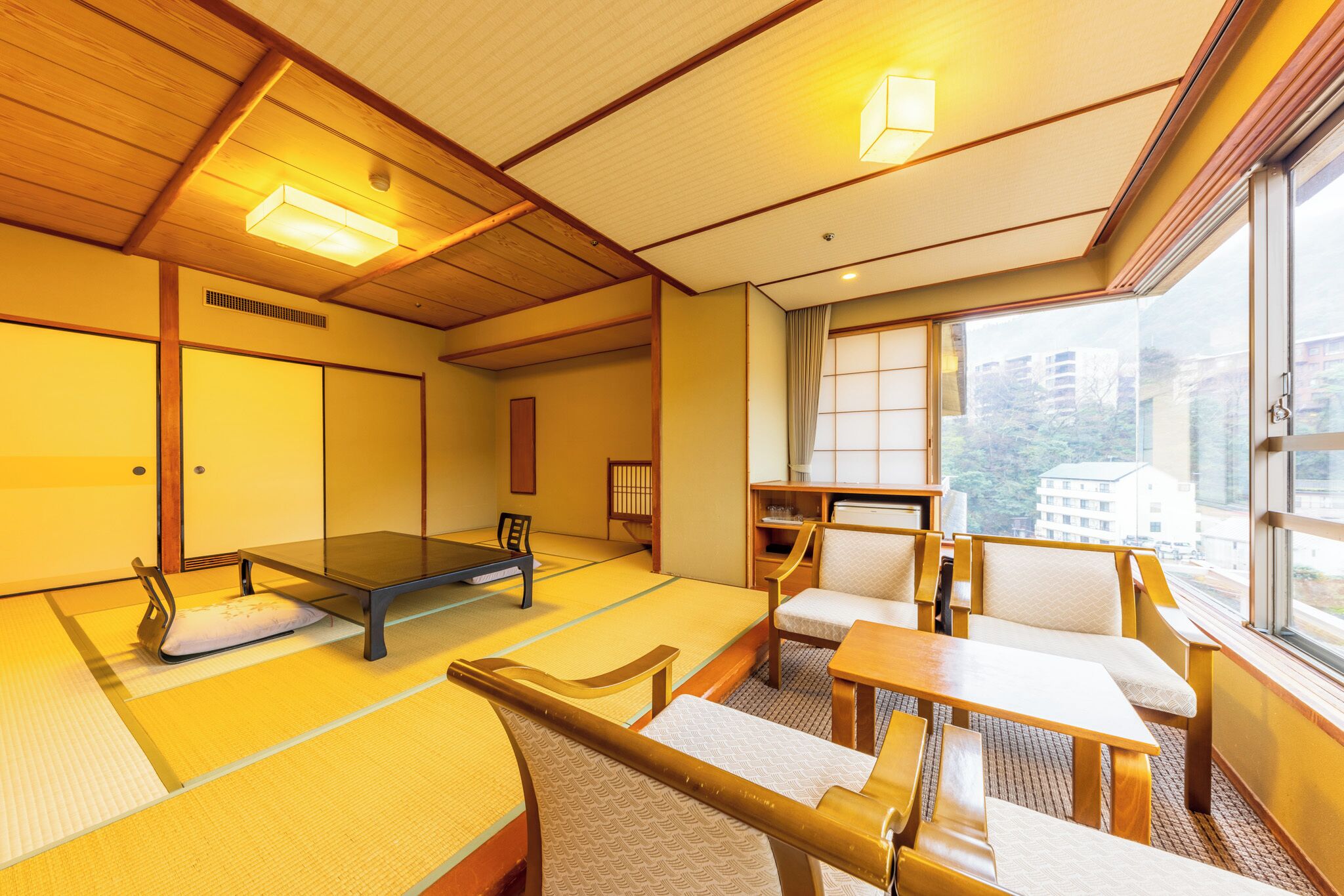 A traditional tatami mat room with a futon bed, tatami seating, and a view of buildings through large windows.