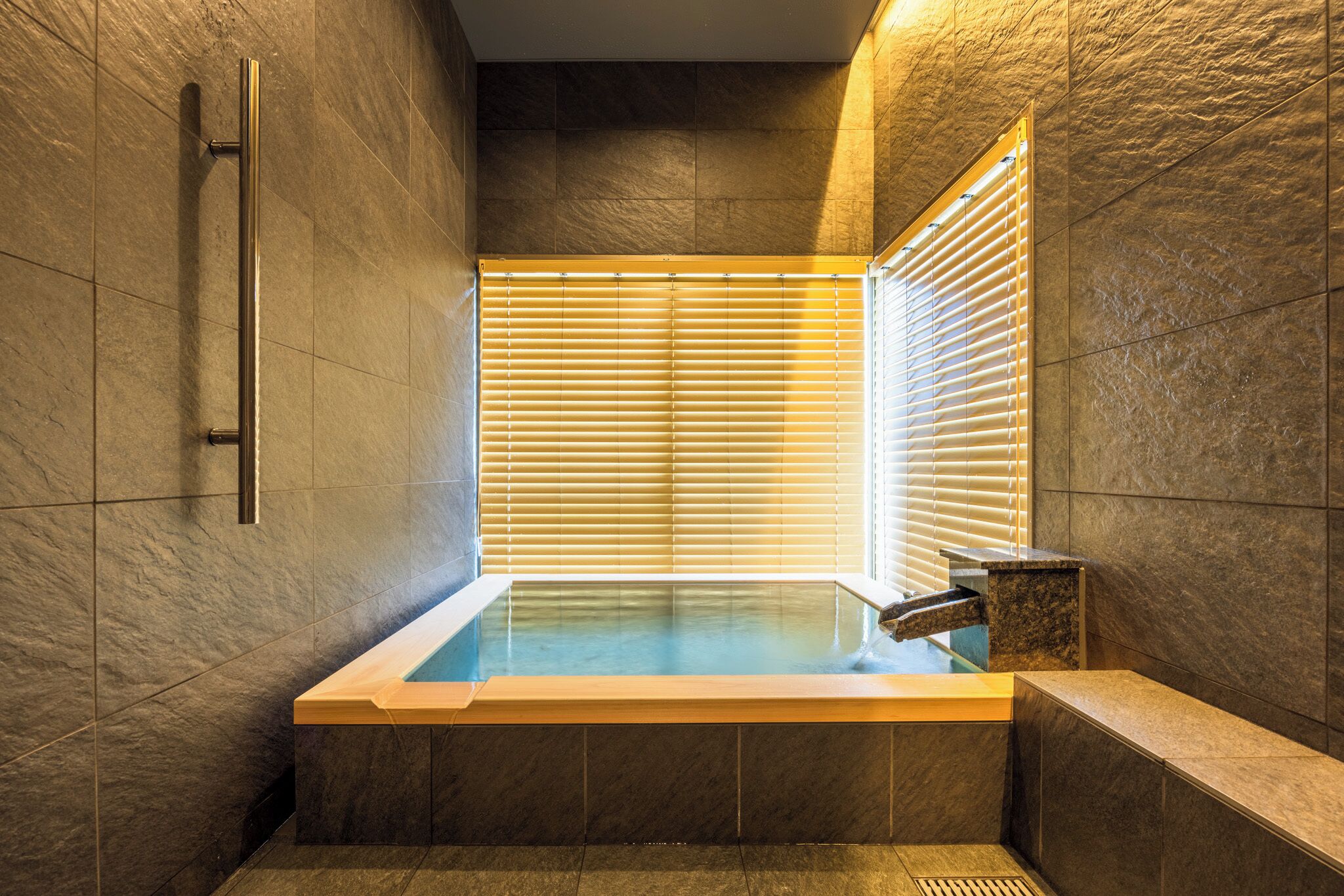 A modern bathroom with a glass-enclosed hot tub, stone walls, and a window with blinds.