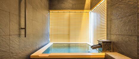 A modern bathroom with a glass-enclosed hot tub, stone walls, and a window with blinds.