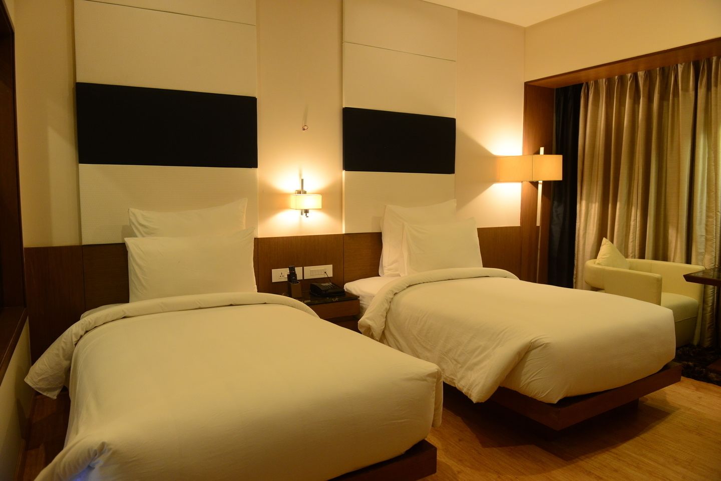 A hotel room with two beds, a nightstand, a lamp, a chair, and a window with curtains.