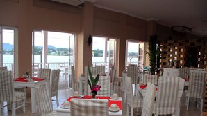 Un comedor con mesas y sillas, ventanas amplias con vistas al agua y montañas, y una pared decorativa con una planta.