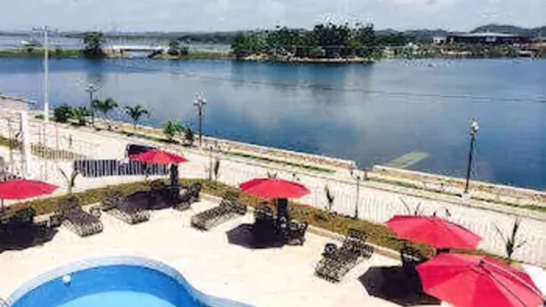 Zona de piscina con sillones de descanso y sombrillas rojas, con vista a una masa de agua y una costa lejana.