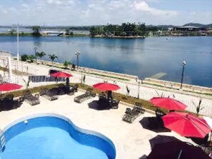 A pool area with lounge chairs and red umbrellas, overlooking a body of water and a distant shoreline. - Ramada Tikal Isla De Flores Hotel (Flores)