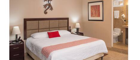 A hotel room with a large bed, two bedside tables with lamps, a red pillow, a framed picture on the wall, and a bathroom visible through an open door.