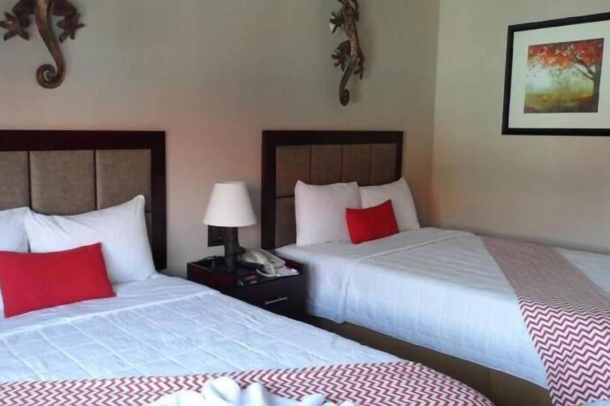 Habitación de hotel con dos camas, cada una con ropa de cama blanca y almohadas rojas, una mesita de noche con una lámpara y un cuadro en la pared.